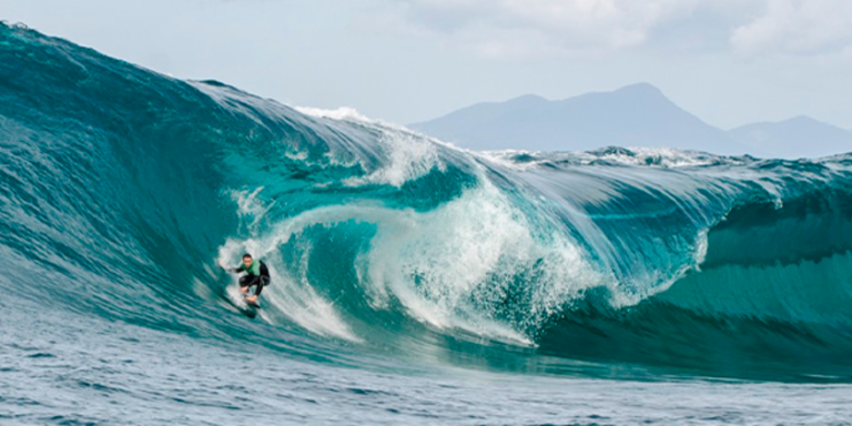 Onda gigante quebra ao fundo de surfista ‘big rider’, formando uma grande espuma branca que persegue o esportista.