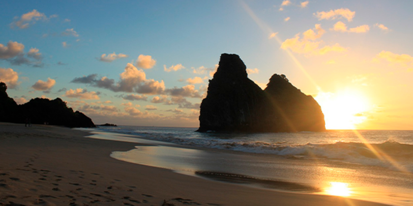 Visão da areia do pôr-do-Sol em meio às pedras na praia Cacimba do Padre, em Fernando de Noronha, com céu claro ao fundo e mar calmo